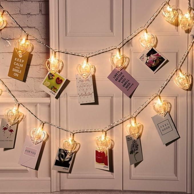 String lights with heart-shaped holders displaying various cards on a white wall.